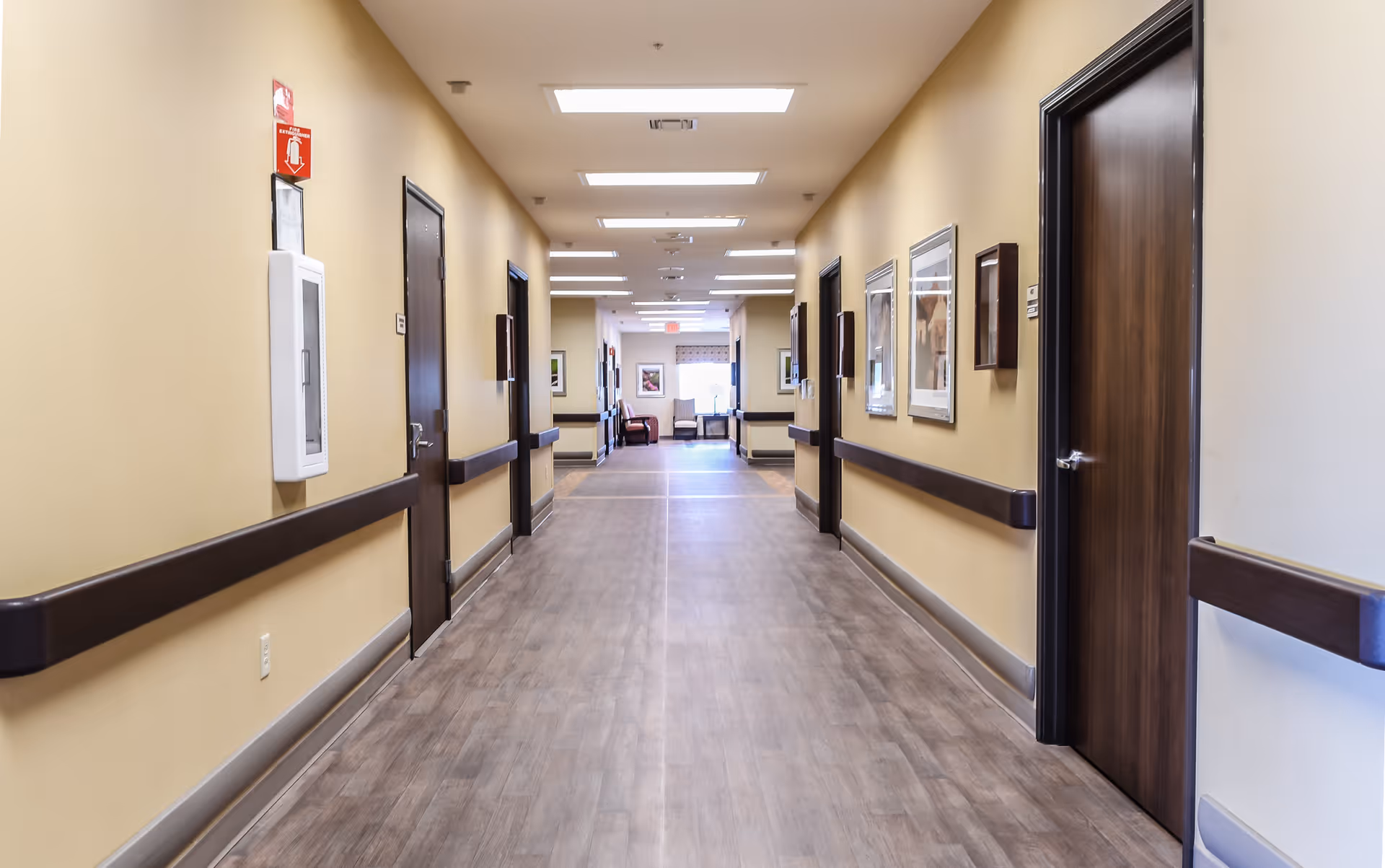A clean, well-lit hallway in a healthcare or rehabilitation facility with beige walls, wooden doors on both sides, handrails along the walls, framed pictures, and a seating area visible at the far end.
