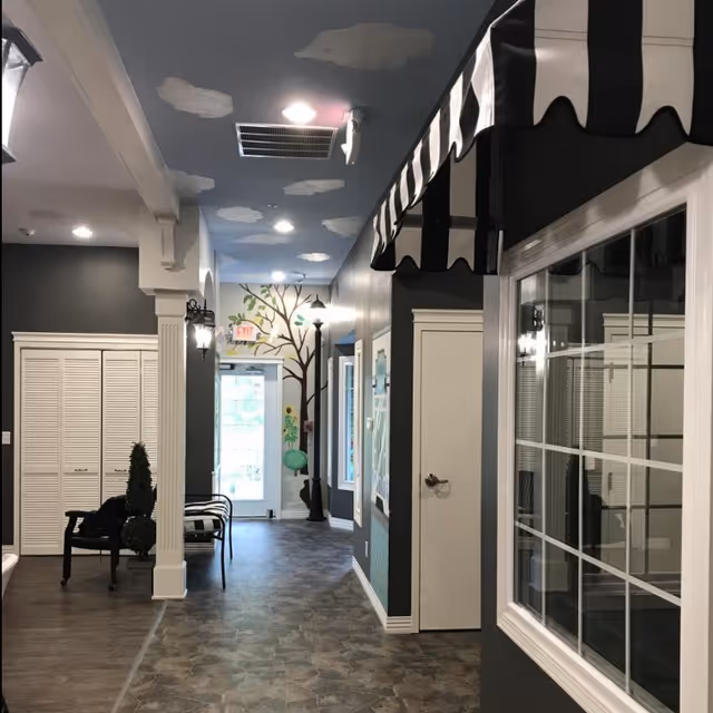 Interior hallway of a senior living facility with dark gray walls, a ceiling painted blue with white clouds, a mural of a tree and flowers near an exit door, black chairs with a small topiary plant, and a window with a black and white striped awning.