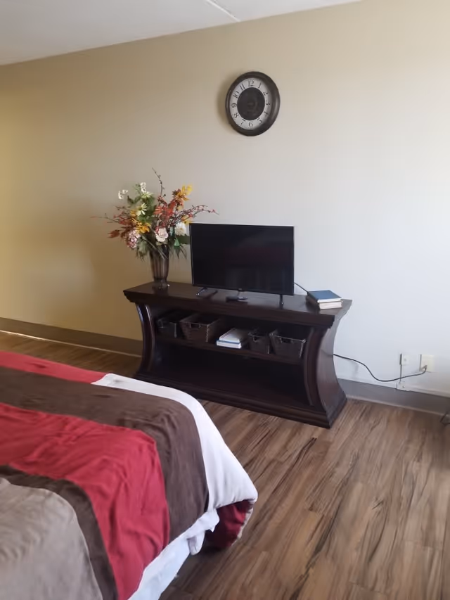 A bedroom corner featuring a bed with a red, brown, and white comforter, a dark wooden TV stand with a flat-screen television, decorative baskets, books, and a vase with artificial flowers. A round wall clock is mounted on the beige wall above the TV stand.