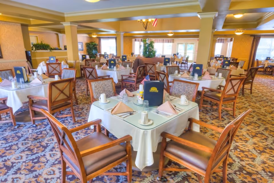 A spacious dining room in The Meadows Senior Living facility with multiple tables covered in white tablecloths, each set with napkins, cups, glasses, and menus. The room features patterned carpet, wooden chairs with cushioned seats, and large windows letting in natural light.