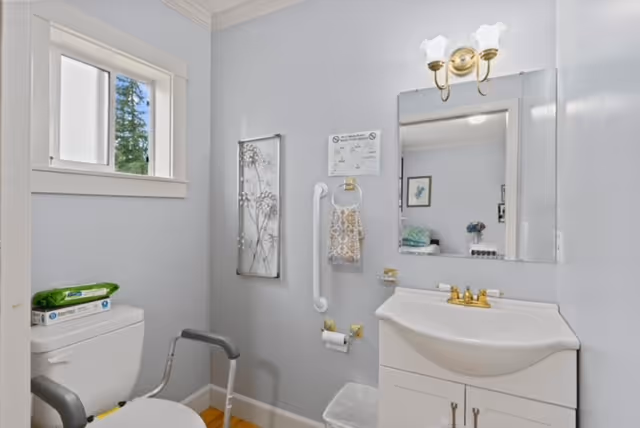 Small bathroom with a toilet (with safety frame), white vanity and mirror, grab bar, and a window.
