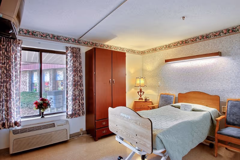 A furnished senior bedroom with an adjustable hospital-style bed, wooden wardrobe and nightstand, chairs, floral curtains, and a window to the outside.