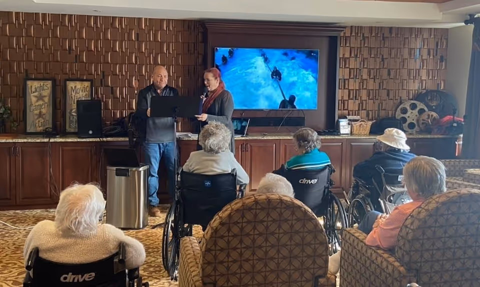 A group of elderly people, some in wheelchairs, seated in a common room watching a presentation by two people standing in front of a large TV screen displaying an underwater scene. The room has wooden cabinets and decorative film reels on the right side.