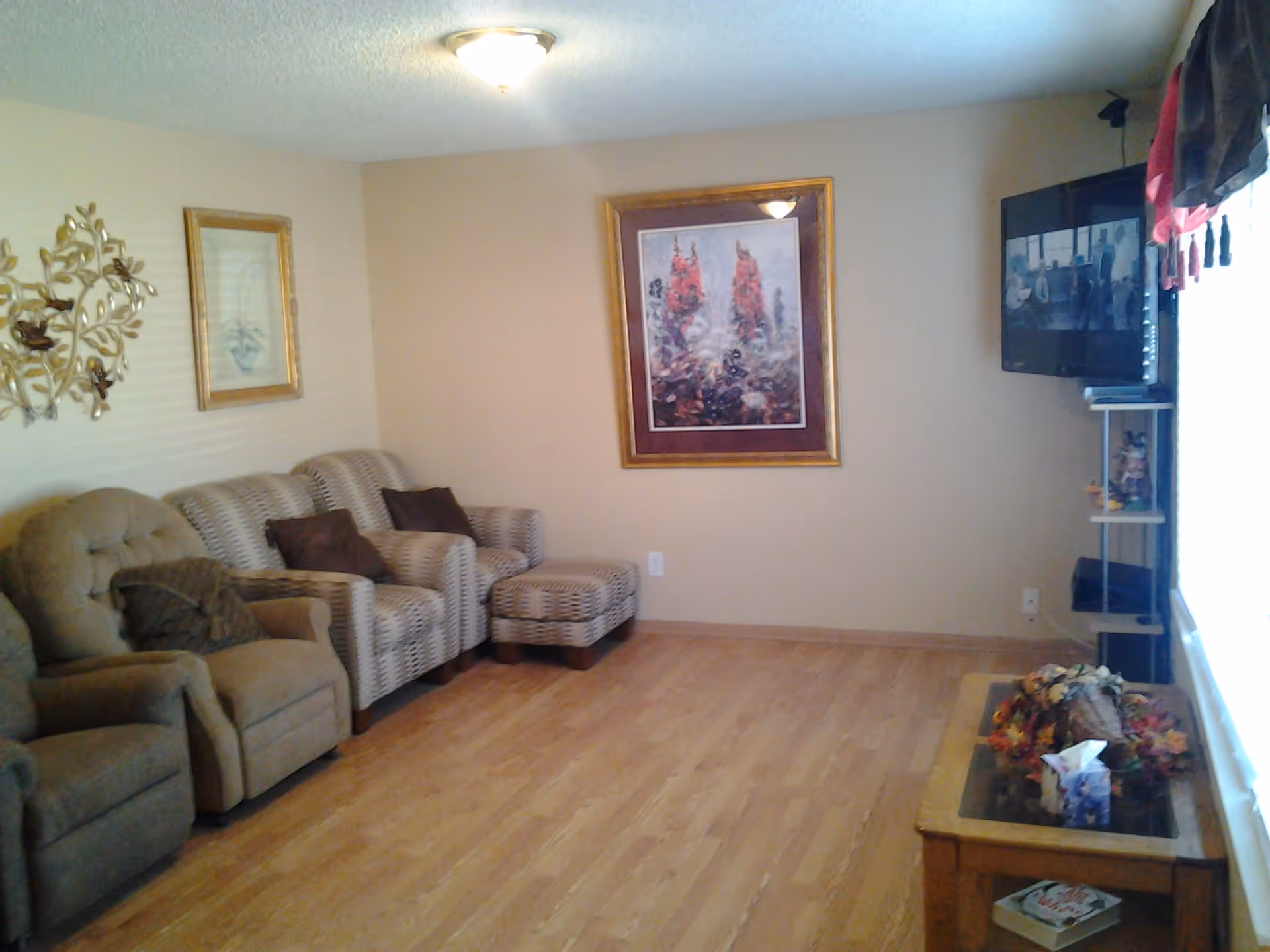 A cozy living room with wooden flooring, featuring a beige recliner chair, a patterned loveseat with matching ottoman, and a wall-mounted flat-screen TV. The walls are decorated with framed artwork and a metal wall sculpture. A wooden coffee table with decorative items and tissues is placed near a large window with curtains.