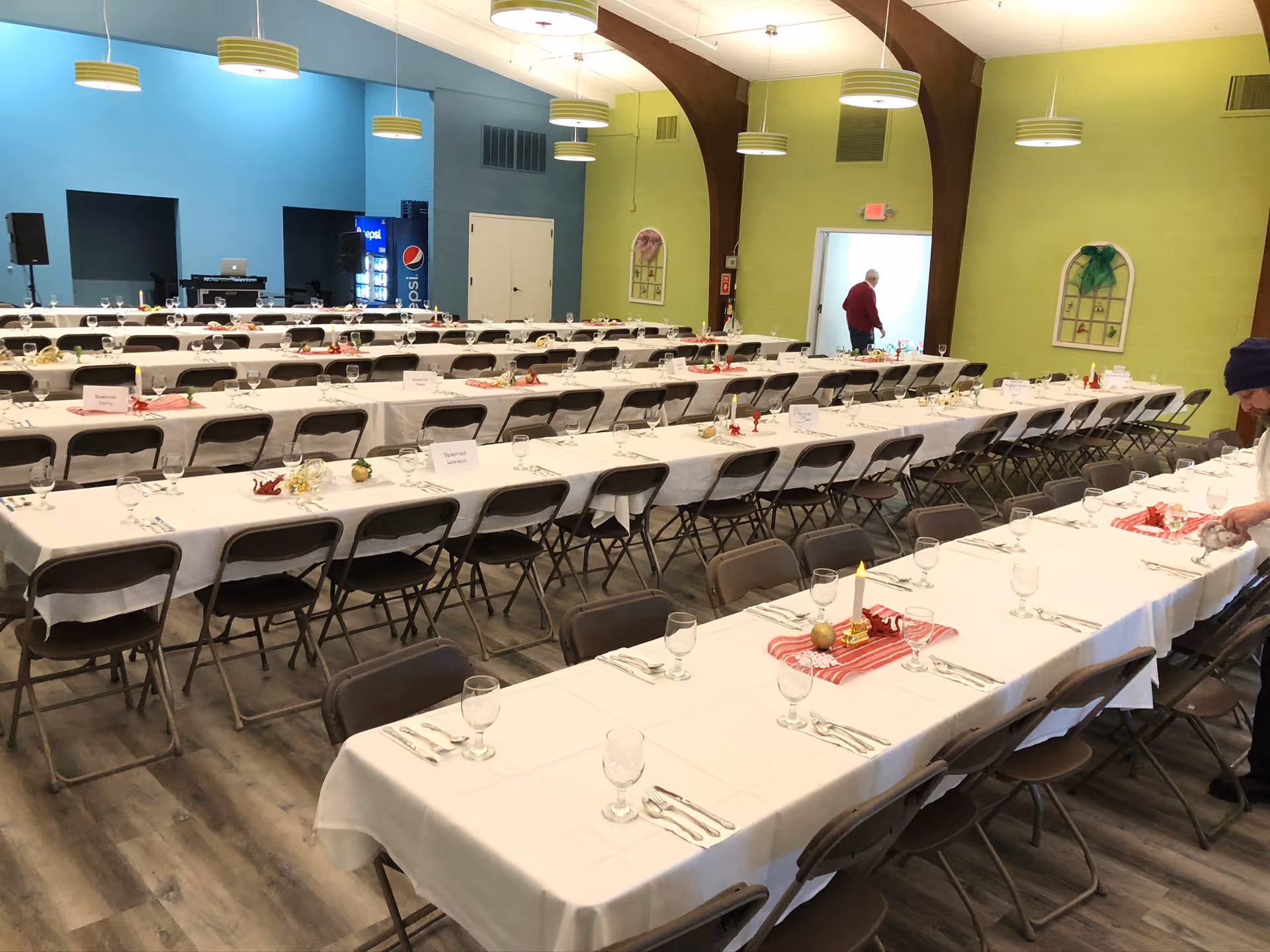 A large dining hall with multiple long tables covered in white tablecloths set with glassware, silverware, and small decorative centerpieces. Folding chairs are arranged along both sides of the tables. The walls are painted blue and green, and there are hanging circular light fixtures from the ceiling. A person is seen near the doorway in the background, and another person is adjusting something on the right side of the image.