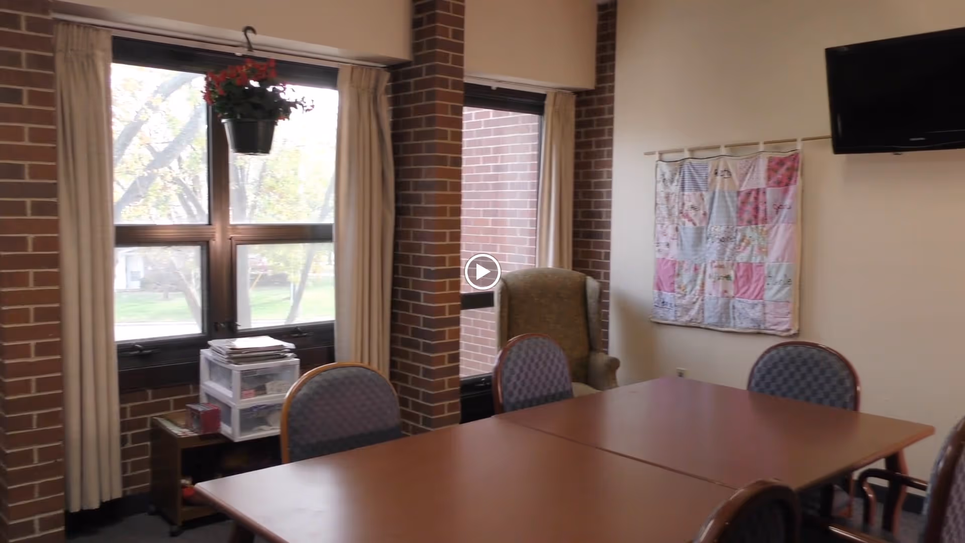 Communal activity/dining room with a large table and chairs, windows with curtains, a wall quilt and a mounted TV.