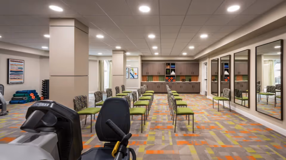 A spacious fitness or activity room with rows of green cushioned chairs arranged in two columns facing a wall with cabinets and shelves. The room features large mirrors on one side, colorful patterned carpet, exercise mats and weights stacked in the corner, and bright overhead lighting.