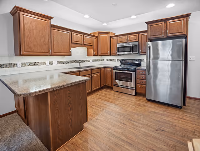 A modern kitchen with wooden cabinets, a stainless steel refrigerator, oven, and microwave. The kitchen features a countertop with a light-colored surface, a tiled backsplash with decorative mosaic accents, and wood-style flooring.