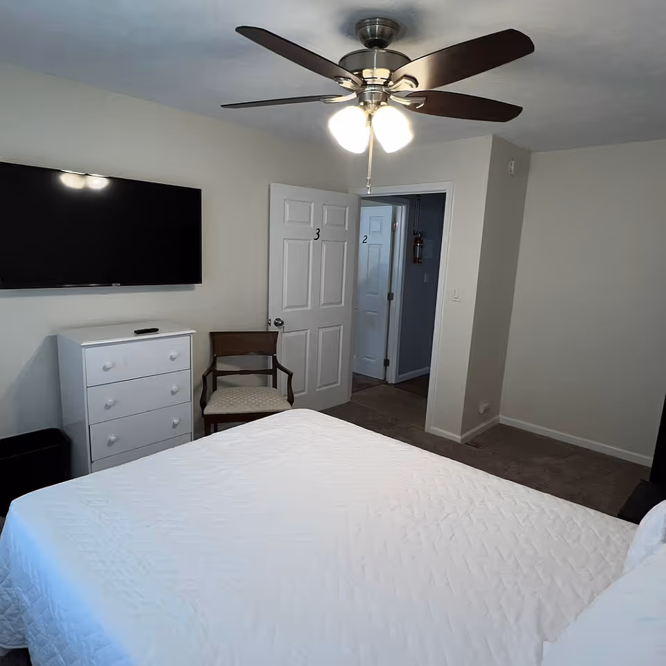 A bedroom with a white bedspread on a bed, a ceiling fan with lights, a wall-mounted flat-screen TV above a white dresser, and a wooden chair. Two doors are visible, one labeled with the number 3 and the other labeled with the number 2.