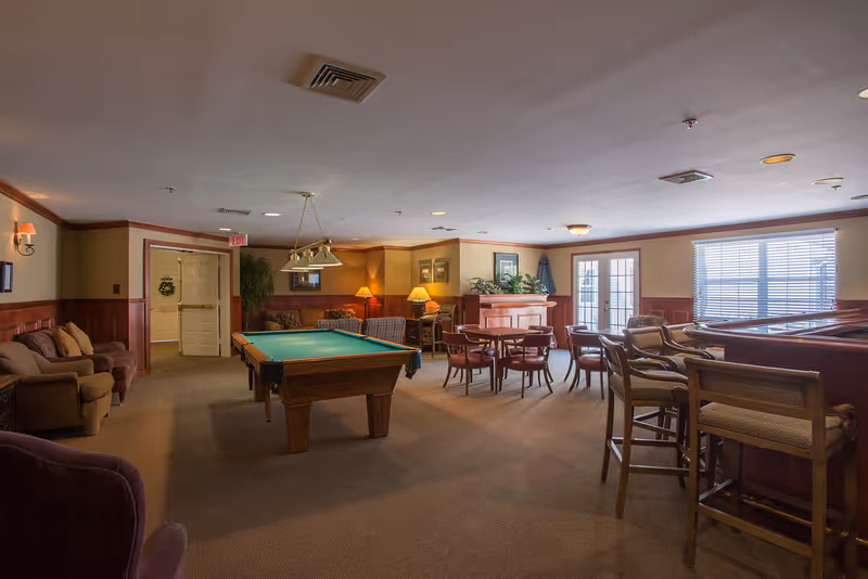 A spacious common room with a pool table in the center, surrounded by various seating options including armchairs, sofas, and tables with chairs. The room has warm lighting, wood paneling on the walls, and large windows letting in natural light.