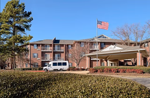 Exterior view of a three-story brick senior living facility with balconies, an American flag on a flagpole, a covered entrance, a white van parked in front, and landscaping including bushes and trees under a clear blue sky.