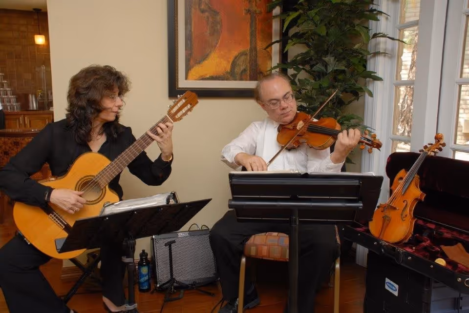 Two musicians playing string instruments indoors; a woman on the left playing a classical guitar and a man on the right playing a violin. They are seated in a room with wooden floors, a large plant, a framed abstract painting on the wall, and a window letting in natural light. A violin case with another violin is open beside the man.