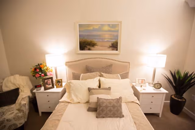 A cozy bedroom with a neatly made bed featuring multiple pillows and a beige headboard. On either side of the bed are white nightstands with table lamps, framed photos, a clock, and a vase of flowers. A framed beach scene painting hangs above the bed. There is a patterned armchair with a throw blanket on the left and a potted plant on the right.