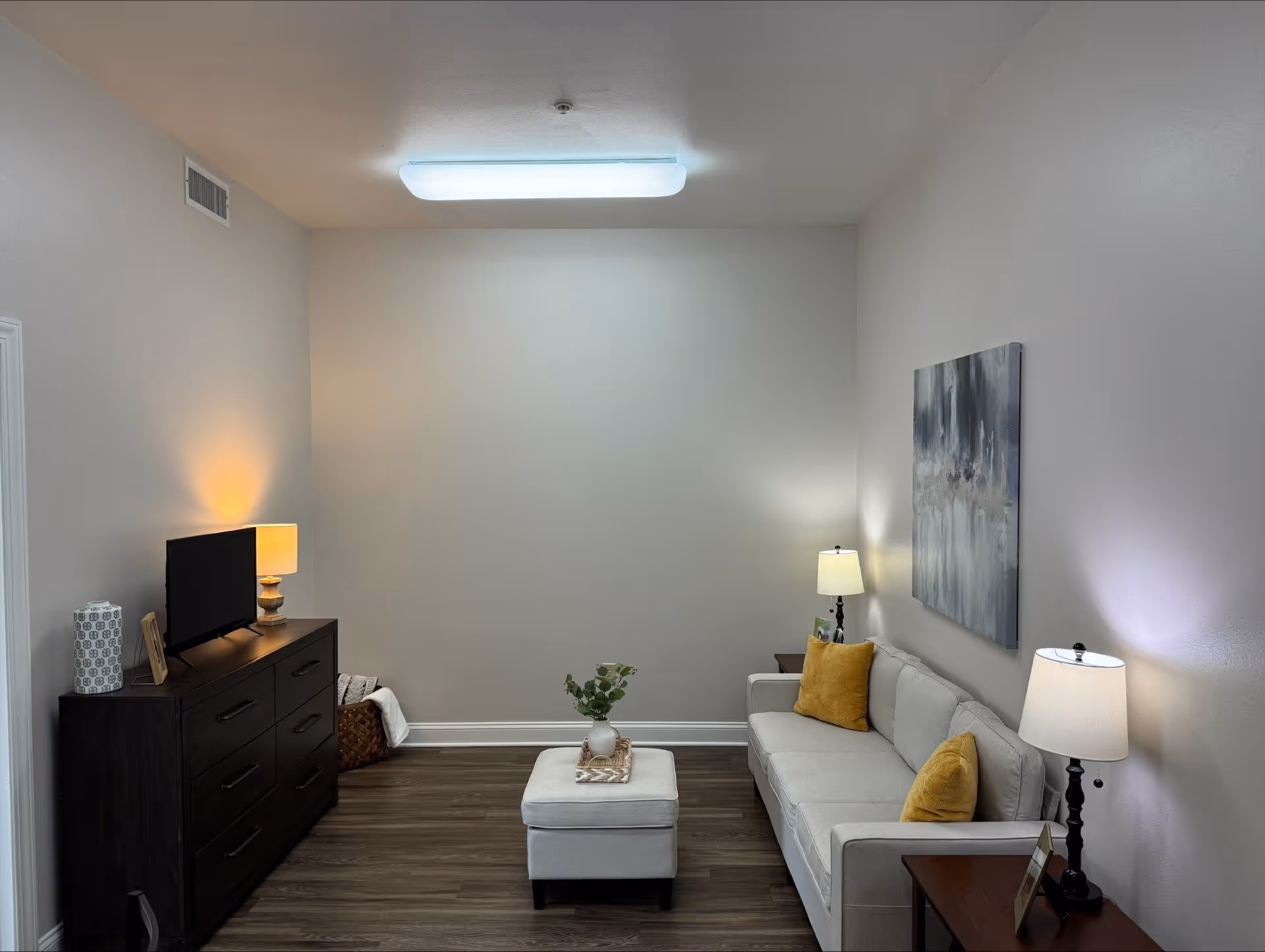 A cozy living room with a light gray sofa adorned with two yellow pillows, a matching ottoman with a small plant on top, two side tables each with a lamp, a dark wooden dresser with a TV and a lamp on it, and a large abstract painting on the wall. The room has wooden flooring and neutral-colored walls.