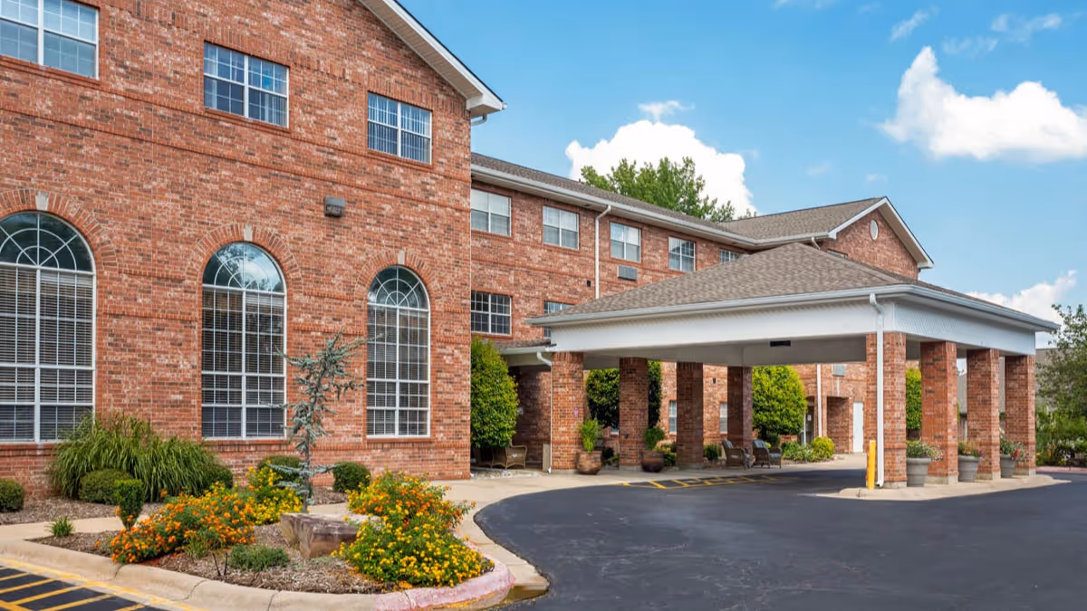 Exterior view of a brick senior living facility building with large arched windows and a covered entrance driveway. There are landscaped flower beds with yellow and orange flowers near the entrance, and a clear blue sky with some clouds in the background.