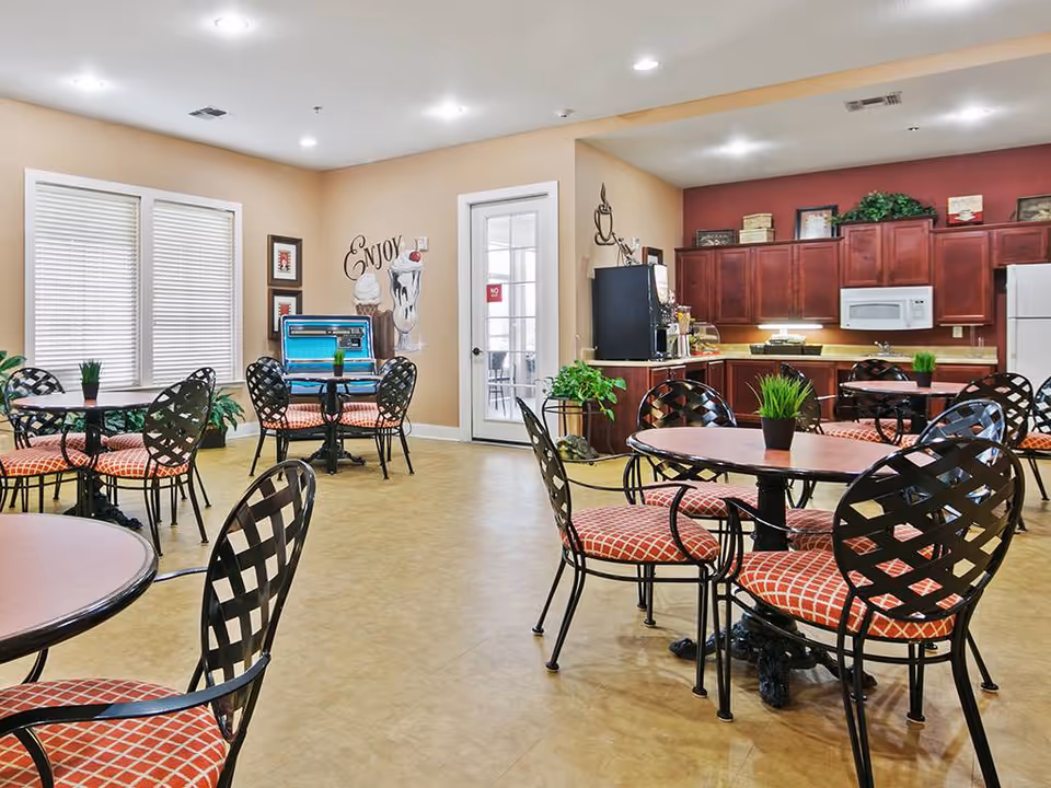 A bright and clean dining area in an assisted living facility with multiple round tables and black metal chairs with red patterned cushions. The room features a kitchenette with wooden cabinets, a microwave, and a refrigerator. There are plants on the tables and around the room, a jukebox against one wall, and a wall decoration with the word 'Enjoy' and images of ice cream sundaes.