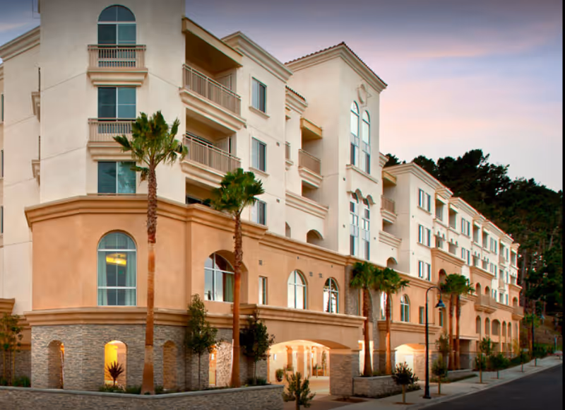 Exterior view of a multi-story senior living facility building named Peninsula Del Rey, featuring beige and light brown walls, multiple balconies, arched windows, palm trees, and a street lamp along the sidewalk at dusk.
