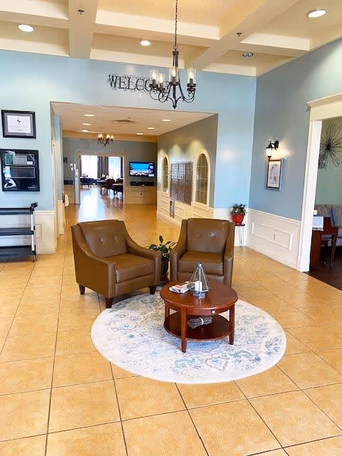 A spacious and well-lit interior common area with two brown leather armchairs arranged around a small round wooden table on a circular rug. The walls are painted light blue with white wainscoting, and there is a chandelier hanging from the ceiling. In the background, a hallway leads to another room with a television mounted on the wall and additional seating visible. Decorative elements include framed pictures, a plant on a small table, and a 'WELCOME' sign above the hallway entrance.