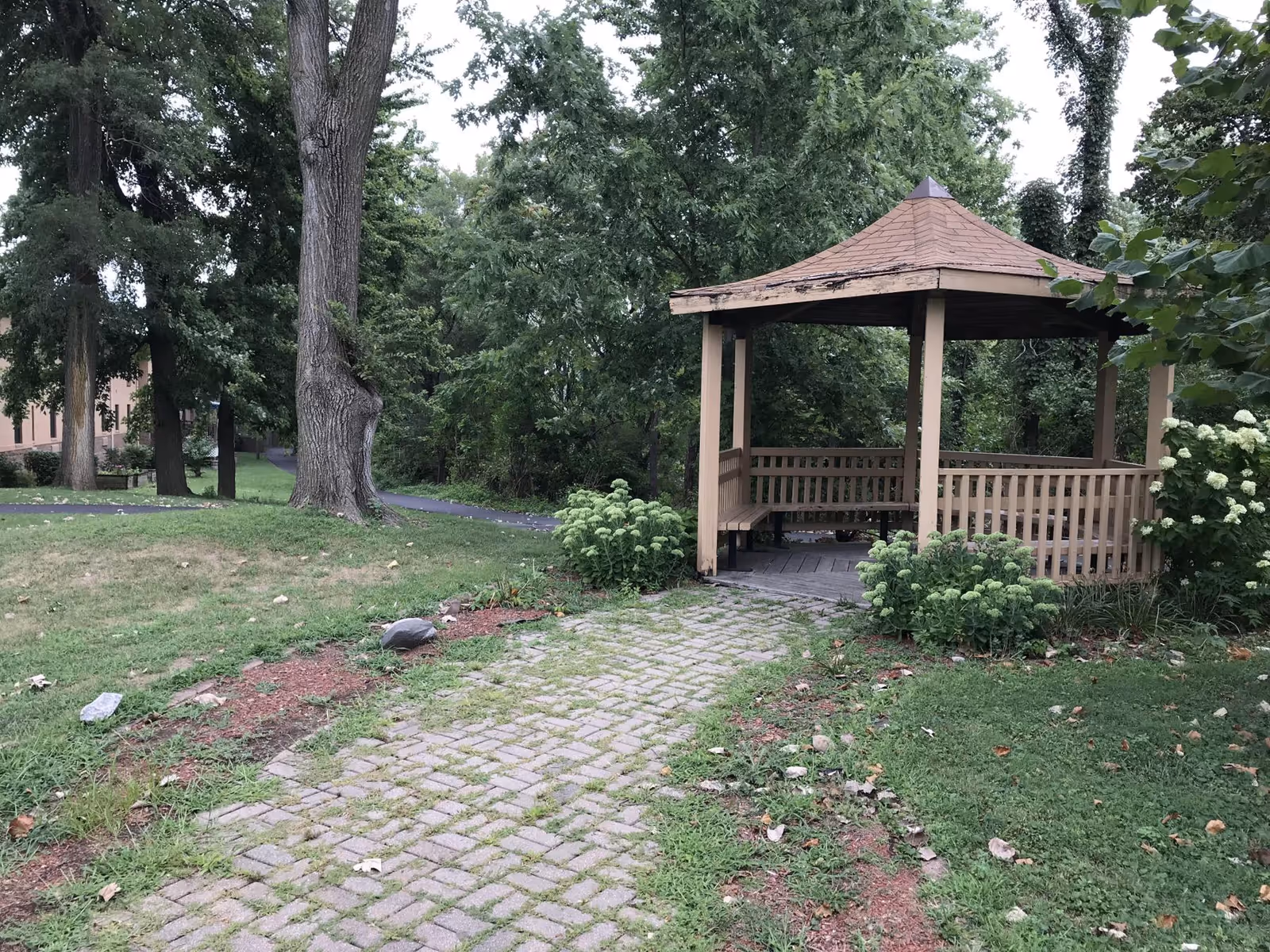 A wooden gazebo with a shingled roof situated in a green outdoor area with trees and bushes. A paved stone pathway leads to the gazebo, and there is a building partially visible on the left side of the image.
