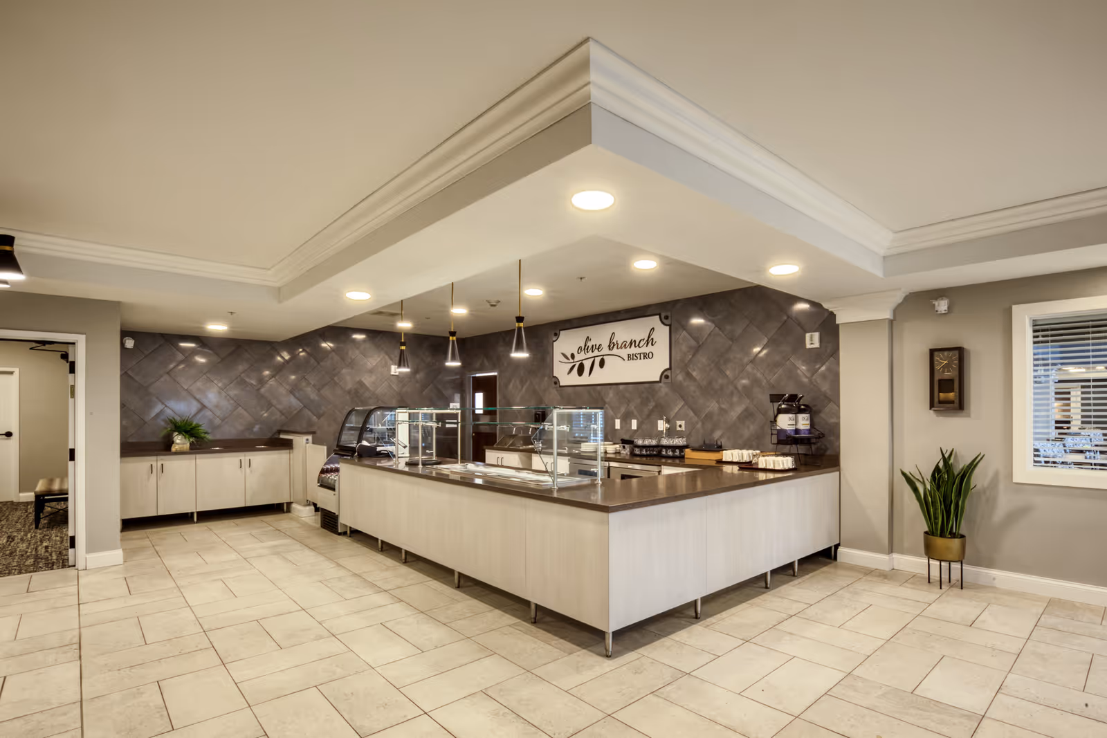 Interior view of a modern dining area named Olive Branch Bistro with a serving counter, glass sneeze guards, pendant lights, and a tiled floor. There is a plant in a pot near a window with blinds and a clock on the wall.