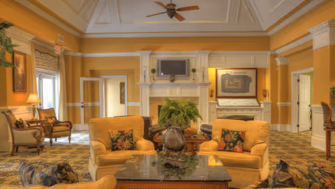 Bright, furnished common lounge with upholstered armchairs and sofas arranged around a coffee table facing a fireplace and wall-mounted TV.