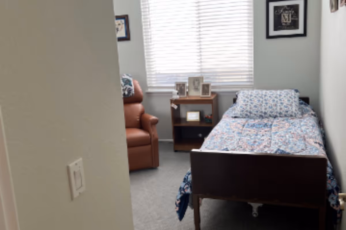 A small bedroom with a single bed covered in a floral patterned quilt and pillow. Next to the bed is a wooden nightstand with framed photos on top. A brown recliner chair is positioned near the window with closed blinds. The walls are light-colored and there are two framed pictures hanging on the walls.