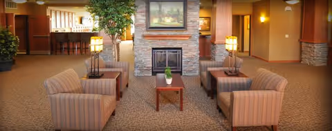 A cozy common area with four striped armchairs arranged around a wooden coffee table with a small plant. The space features a stone fireplace with a framed landscape painting above it, two table lamps on side tables, and a large indoor plant. The background shows a bar counter with stools and warm lighting.