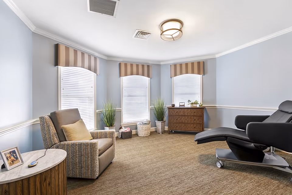 A cozy room with light blue walls and three large windows with striped valances. The room features a comfortable armchair with a yellow pillow, a small round wooden side table with a framed photo and a remote control, two potted plants, a wooden dresser, and a black reclining chair on wheels. The carpet is beige, and the ceiling has a modern light fixture.