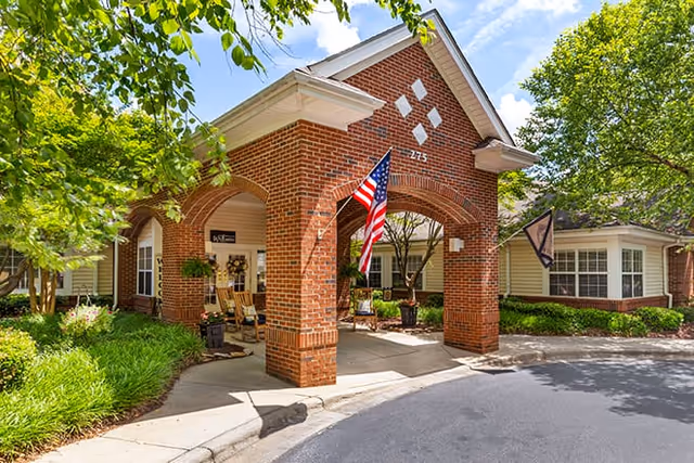 Entrance of a senior living facility with a brick archway, an American flag hanging from the arch, and a covered driveway. There are rocking chairs and potted plants under the archway, surrounded by green trees and shrubs on a sunny day.