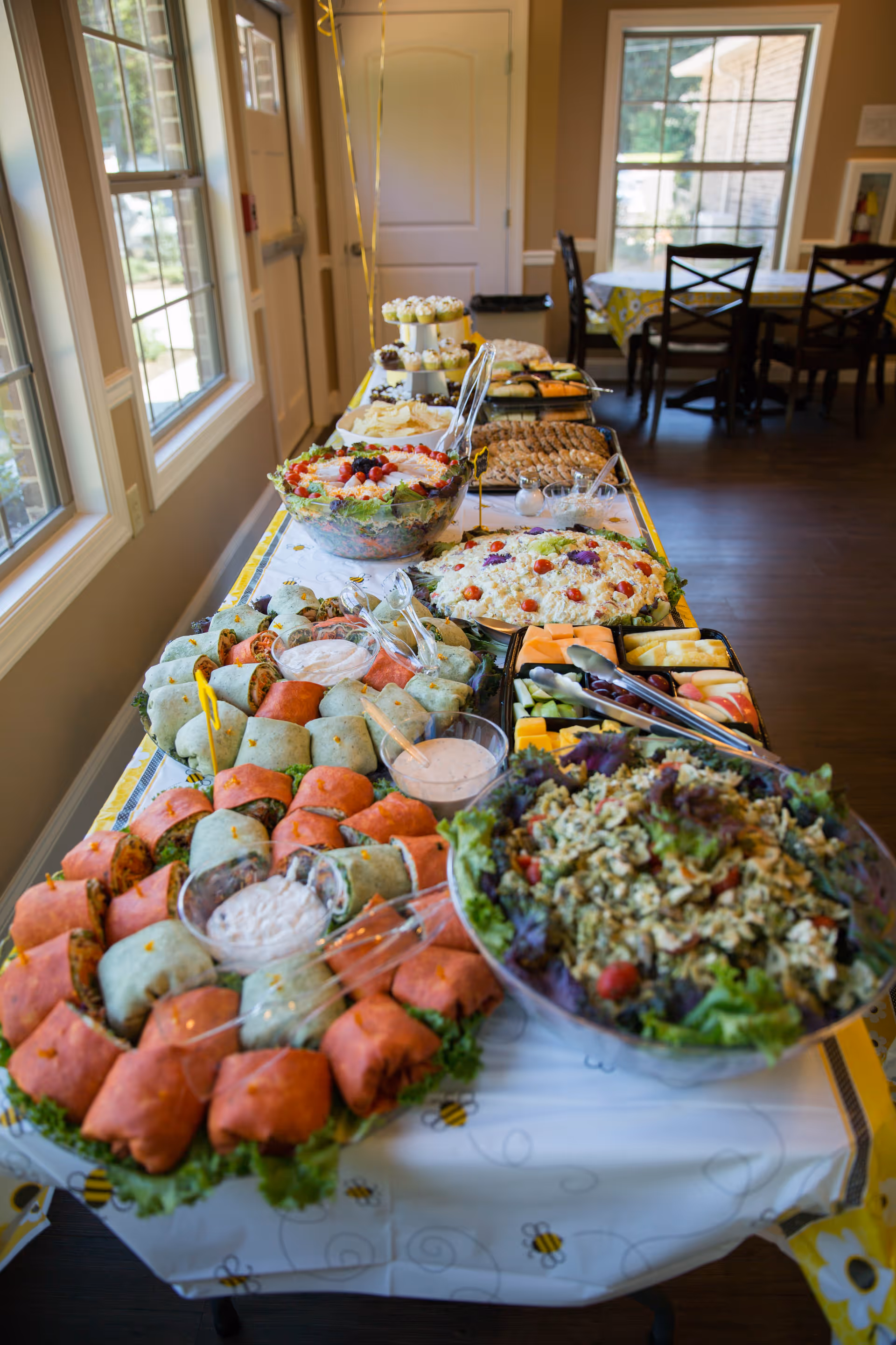 A long table covered with a white tablecloth decorated with bees, set up with a variety of food including wraps, salads, fruit trays, and desserts in a bright room with large windows and wooden floors.