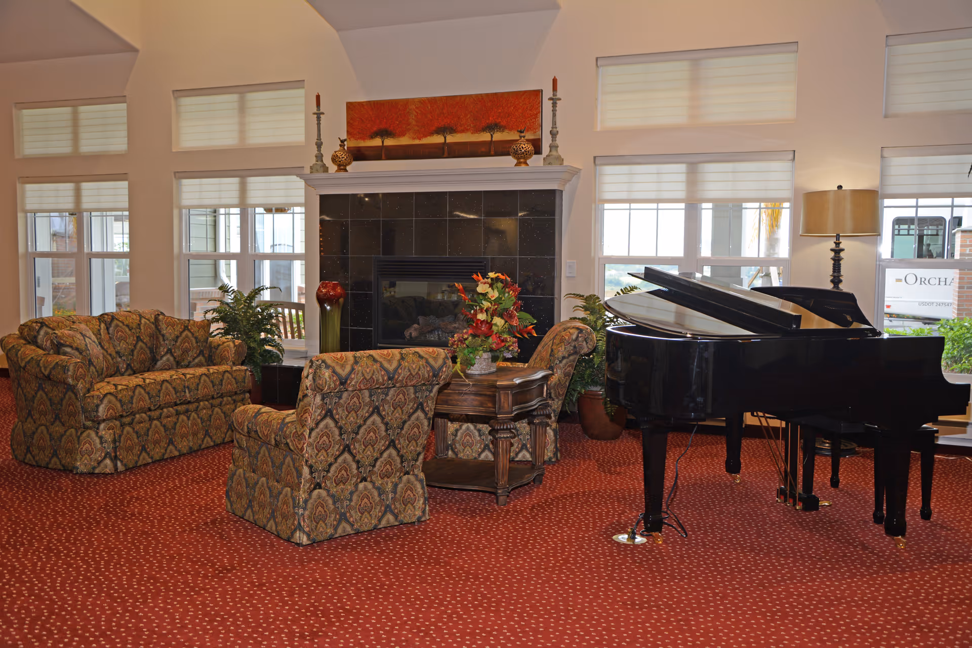 A cozy living room area with patterned upholstered chairs and a sofa arranged around a wooden coffee table with a floral centerpiece. Behind the seating is a black tiled fireplace with decorative items and a painting above it. To the right, there is a black grand piano and a floor lamp near large windows with white blinds, letting in natural light.