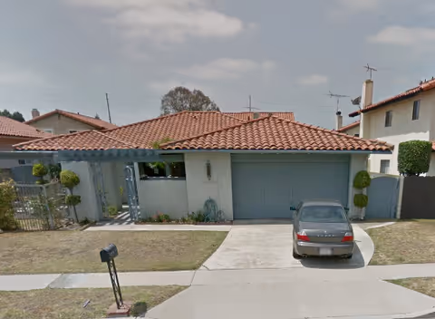 Single-story house with a red tiled roof and a gray garage door. A gray car is parked in the driveway. The front yard has a small lawn and trimmed bushes. The sky is partly cloudy.