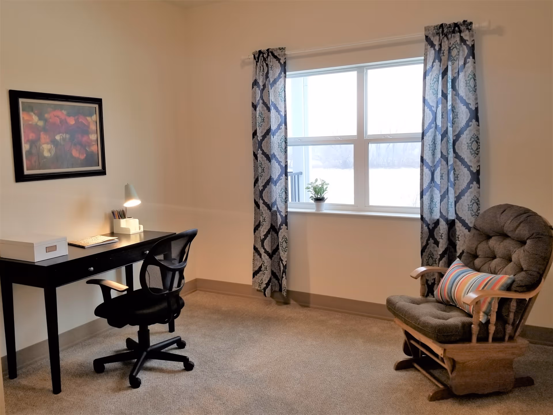 Sunlit room with a desk and office chair on the left and a cushioned rocking chair by a window with patterned curtains.