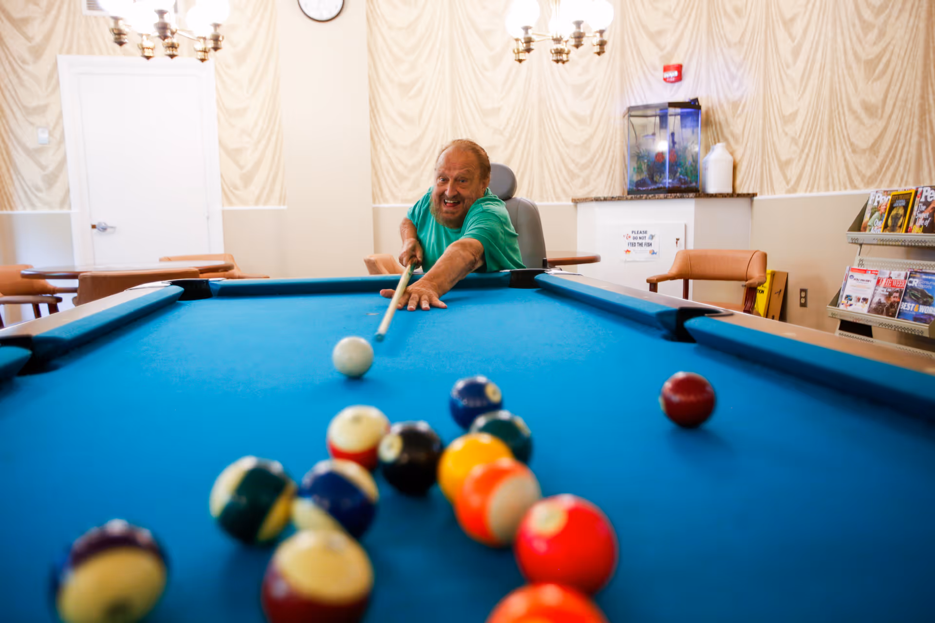 An elderly man in a green shirt playing pool in a recreational room with a blue pool table, chairs, a fish tank, and a magazine rack in the background.