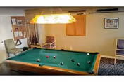 A room with a green felt pool table in the center, surrounded by chairs and a bulletin board on the wall. A rectangular light fixture hangs above the pool table, illuminating the playing surface.