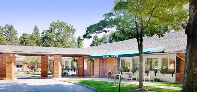 Front entrance of a single-story brick senior living facility with a covered walkway, green awning, benches, and trees.