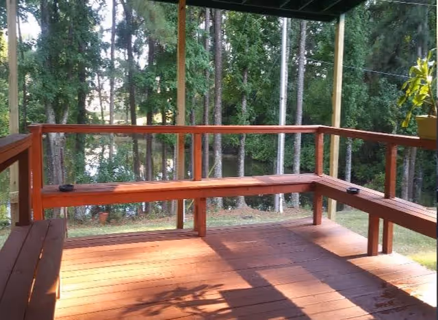 Covered wooden deck with built-in bench seating overlooking a wooded yard with a glimpse of water.