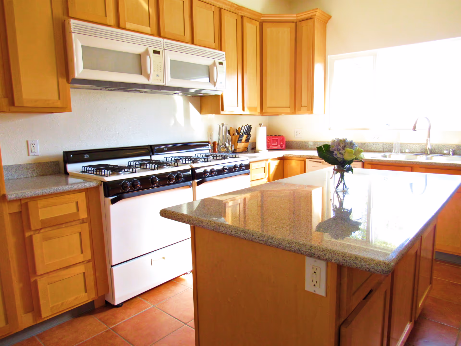 Bright kitchen with light wood cabinets, a large granite island with a vase of flowers, and twin gas ranges under a microwave.