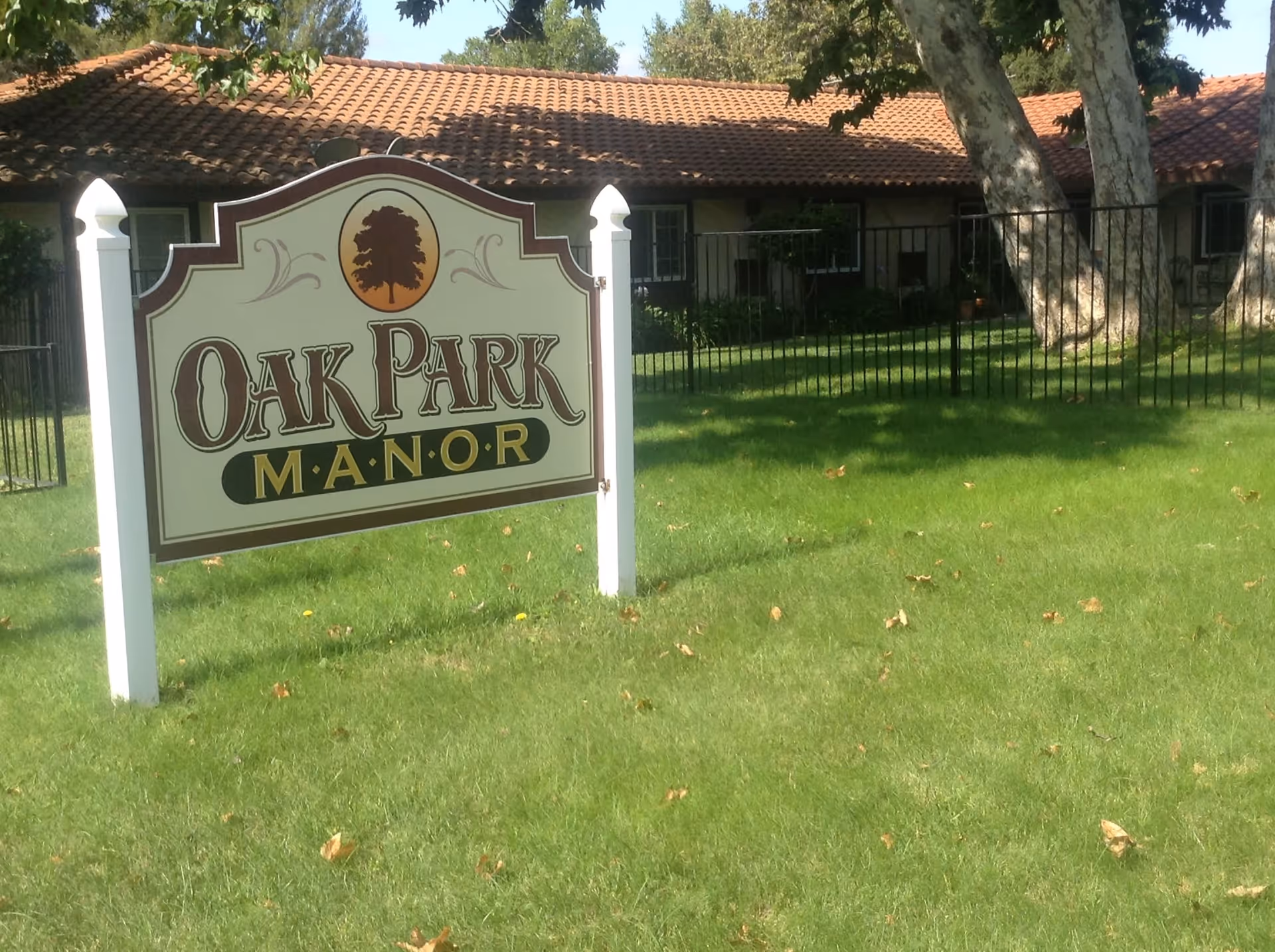A green lawn with a white sign that reads 'Oak Park Manor' in front of a building with a tiled roof and several windows, surrounded by trees and a black metal fence.