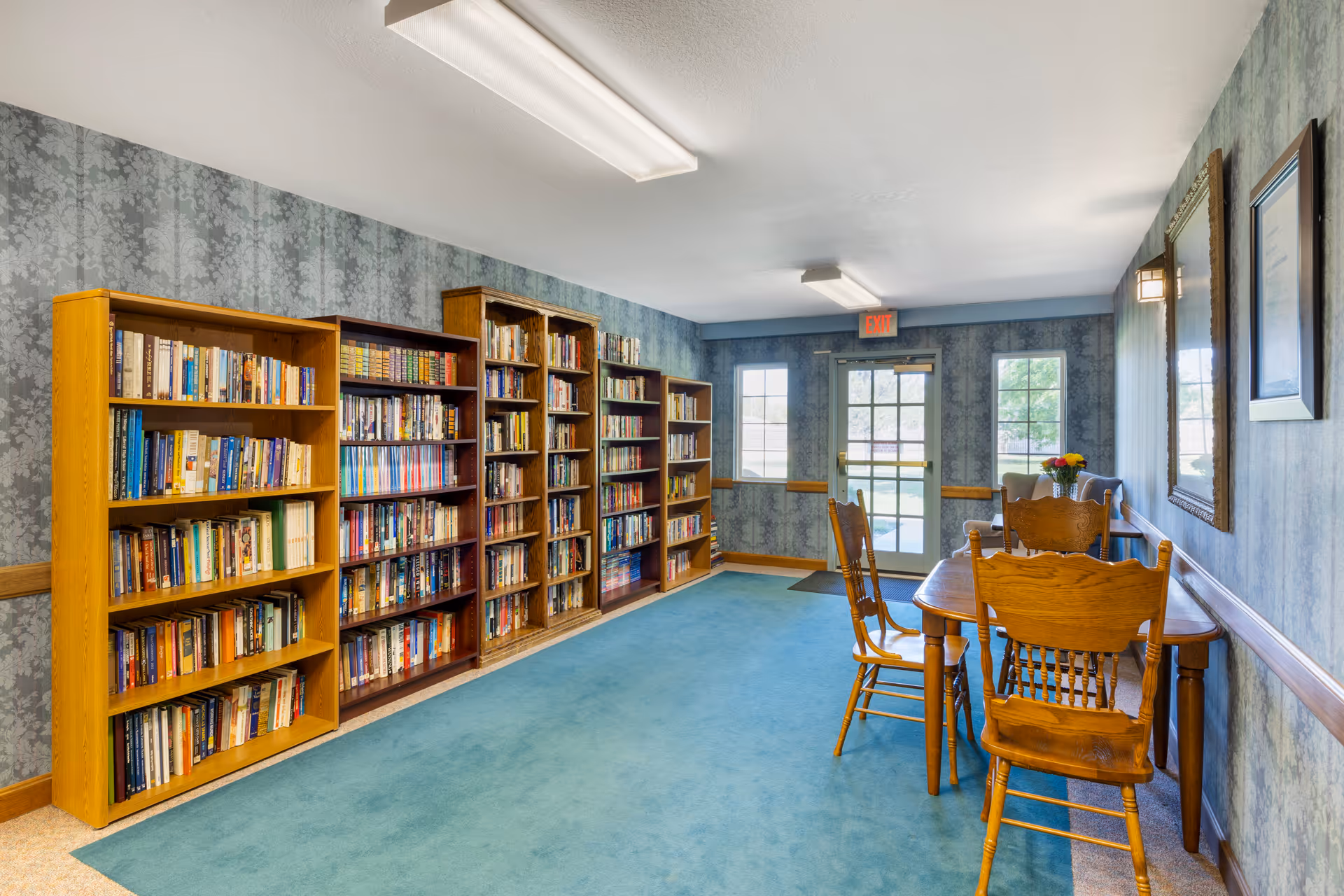 A quiet library room with several wooden bookshelves filled with books along the left wall. On the right side, there is a wooden table with four wooden chairs. The room has blue patterned wallpaper, blue carpet, and fluorescent ceiling lights. At the far end, there is a glass door and two windows letting in natural light.