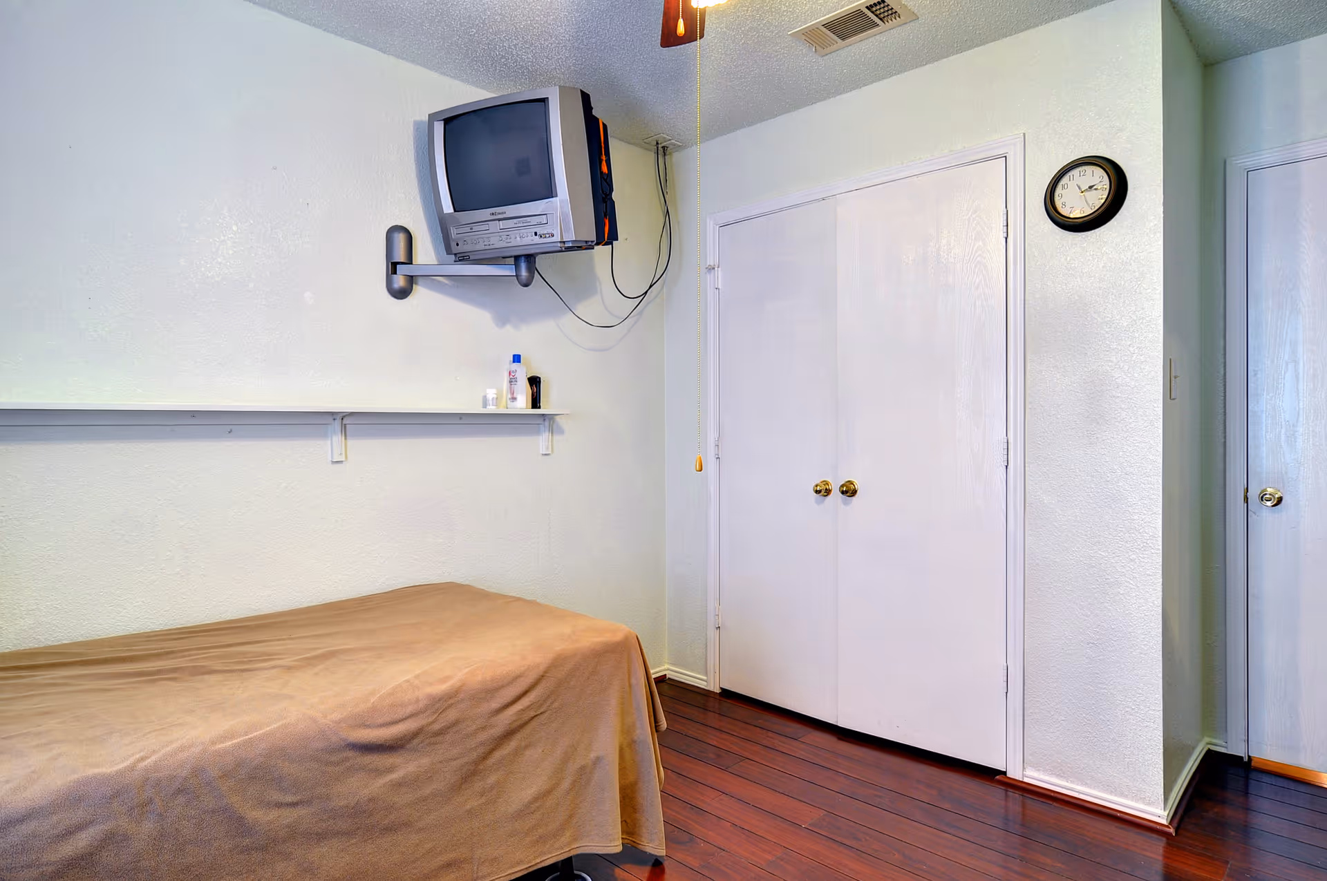 Small bedroom with a single bed covered by a brown blanket, a wall-mounted CRT TV above a shelf with toiletries, double closet doors, and a wall clock.