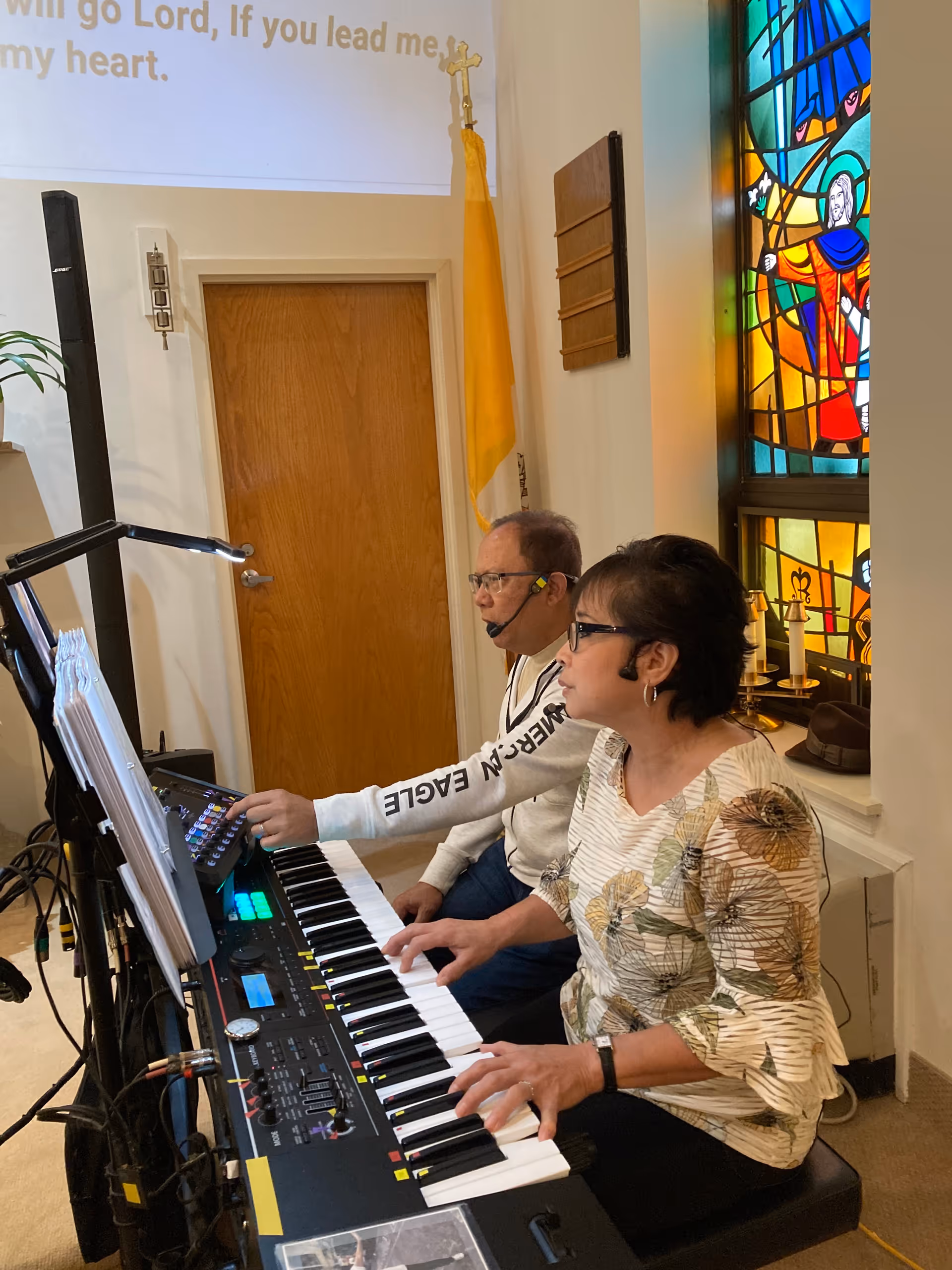 Two people playing an electronic keyboard in a room with a stained glass window and a wooden door. One person is wearing a headset microphone and a white sweatshirt, while the other is wearing glasses and a floral patterned blouse. There is a yellow and white flag with a cross on a stand near the door.