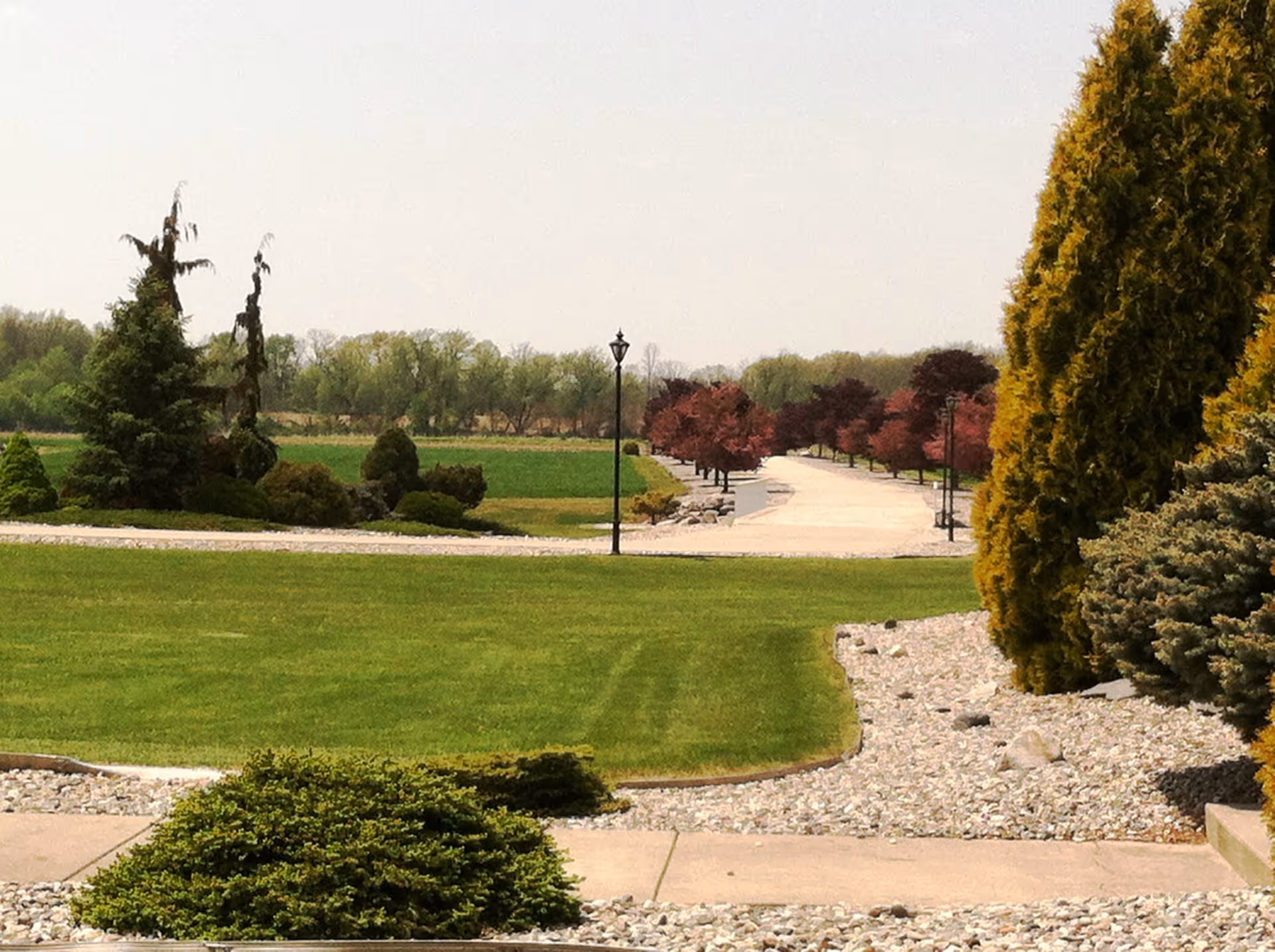 A landscaped outdoor area with a green lawn, various bushes and trees, a paved walkway lined with red-leaved trees, and a lamppost in the center. The sky is clear and the scene appears calm and well-maintained.