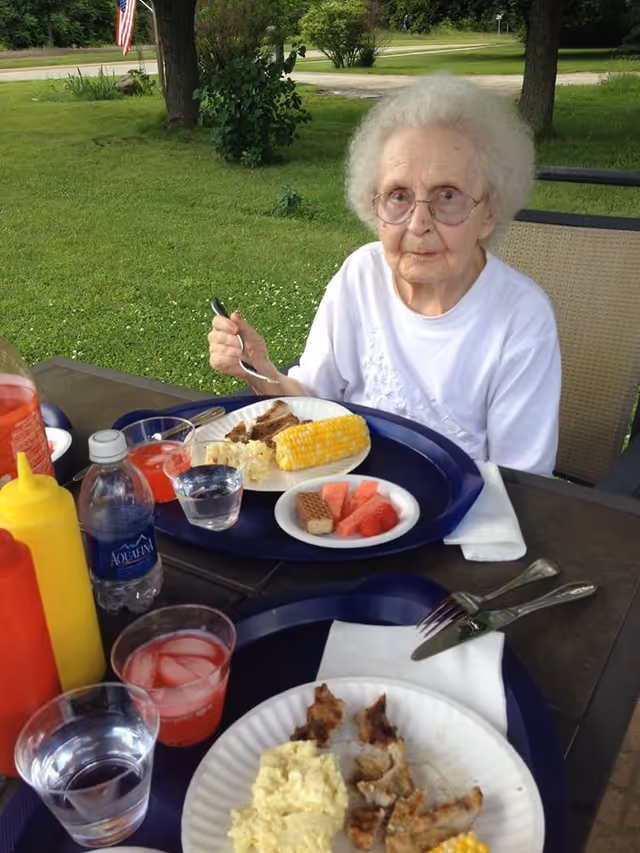 An elderly woman with white hair and glasses sitting outdoors at a table with a meal in front of her. The meal includes corn on the cob, watermelon, strawberries, a wafer cookie, and some meat. There are condiments, a water bottle, and drinks on the table. The setting is a grassy area with trees and an American flag visible in the background.