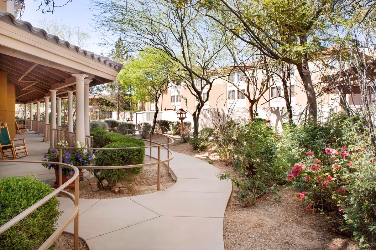 Outdoor garden area at Sunrise of Scottsdale featuring a paved walkway with handrails, surrounded by green bushes, flowering plants, and trees. A covered porch with rocking chairs is visible on the left side, and a multi-story building is in the background under a clear blue sky.