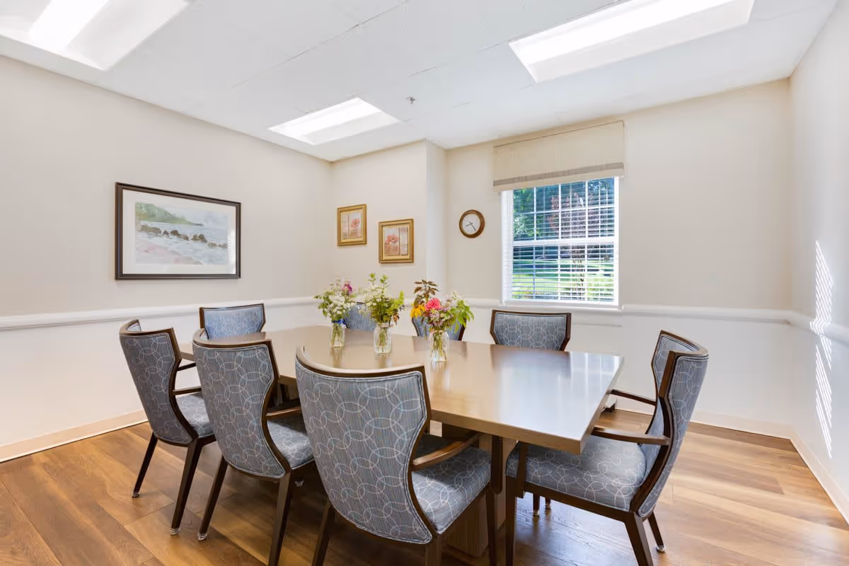 A bright dining room with a rectangular wooden table surrounded by eight upholstered chairs with patterned fabric. Three small vases with fresh flowers are placed on the table. The room has light-colored walls with three framed pictures and a clock on the wall. A window with blinds lets in natural light, and the floor is wood.