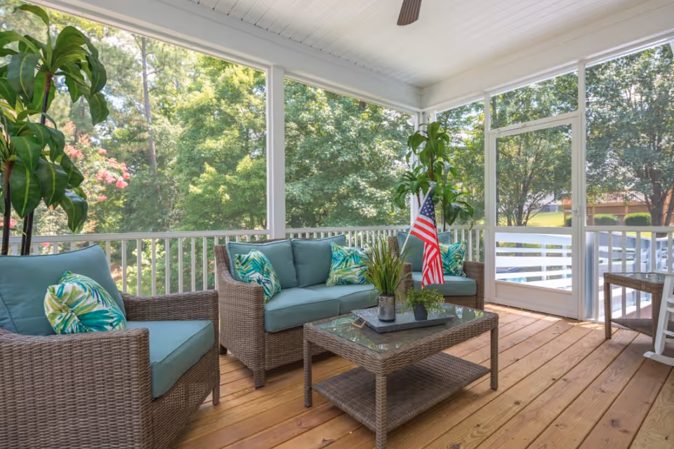 A bright and airy screened-in porch with wicker furniture including a sofa and two chairs with blue cushions and tropical leaf patterned pillows. A glass-top wicker coffee table holds a tray with potted plants and an American flag. The porch overlooks a lush green garden with trees and shrubs.