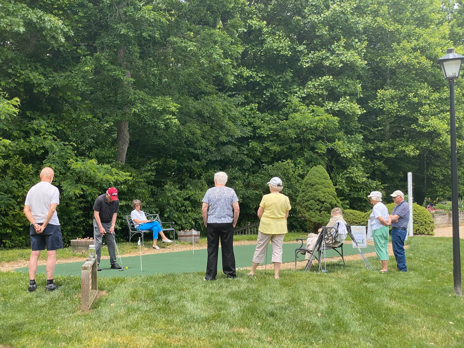 A group of elderly people outdoors at Ardenwoods Retirement Community, some standing and some sitting on benches, watching a man in a red cap putting a golf ball on a small green putting area surrounded by trees and bushes.
