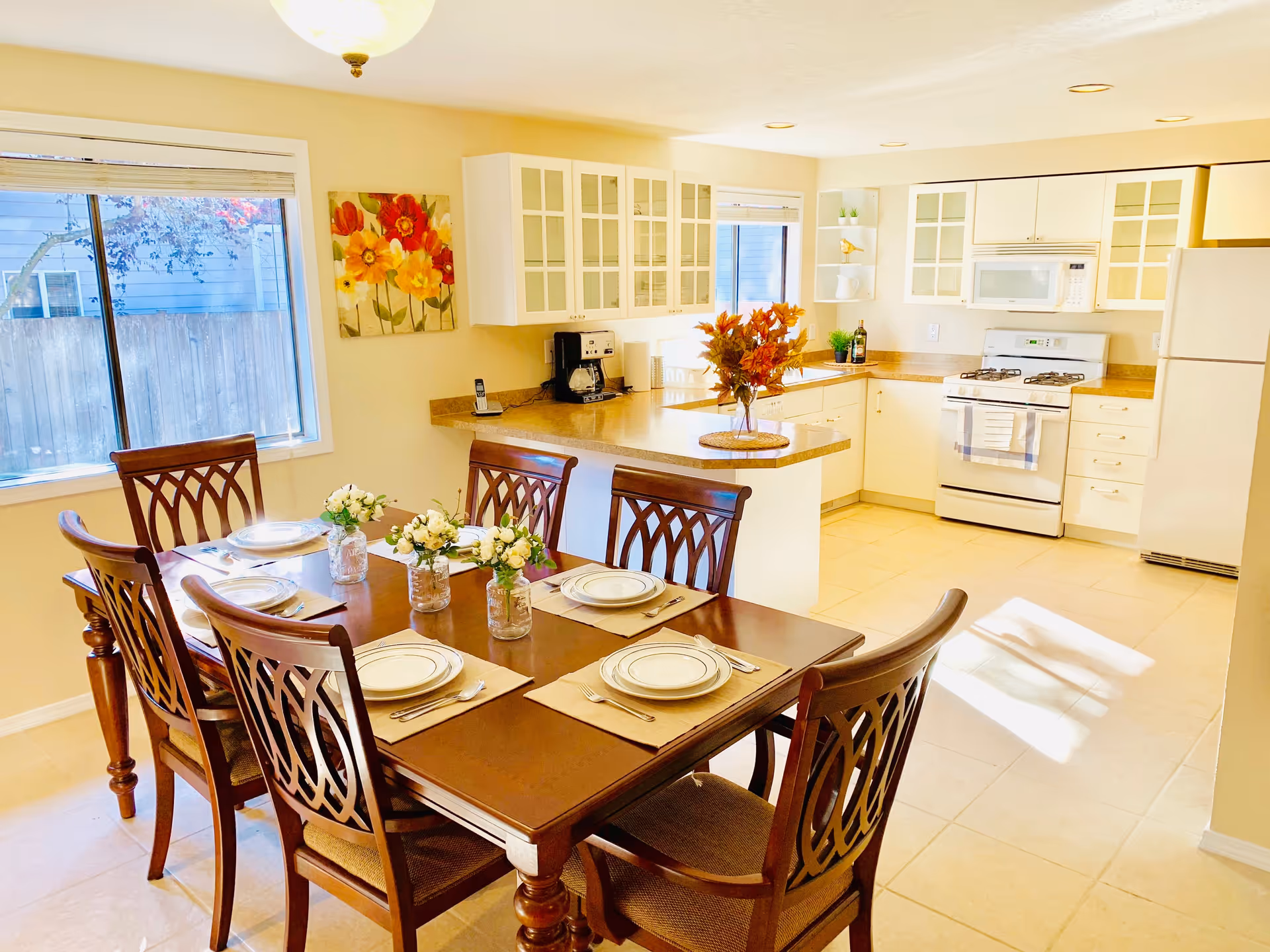 Bright kitchen and dining area with a wooden dining table set for six people, decorated with small flower arrangements. The kitchen features white cabinets with glass doors, a white refrigerator, stove, microwave, and a countertop island with a vase of autumn leaves. A large window allows natural light to fill the space.