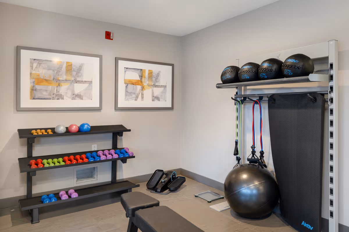 A small fitness room with a rack of colorful dumbbells, exercise balls, resistance bands, a yoga mat, and a bench. Two abstract paintings hang on the wall above the dumbbell rack.